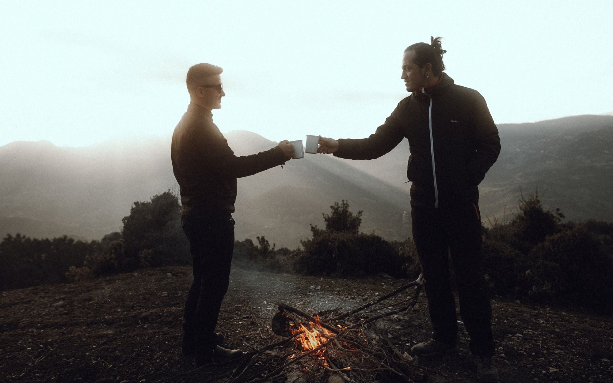 Two men enjoying a sunset with mugs by a campfire in a rural setting
